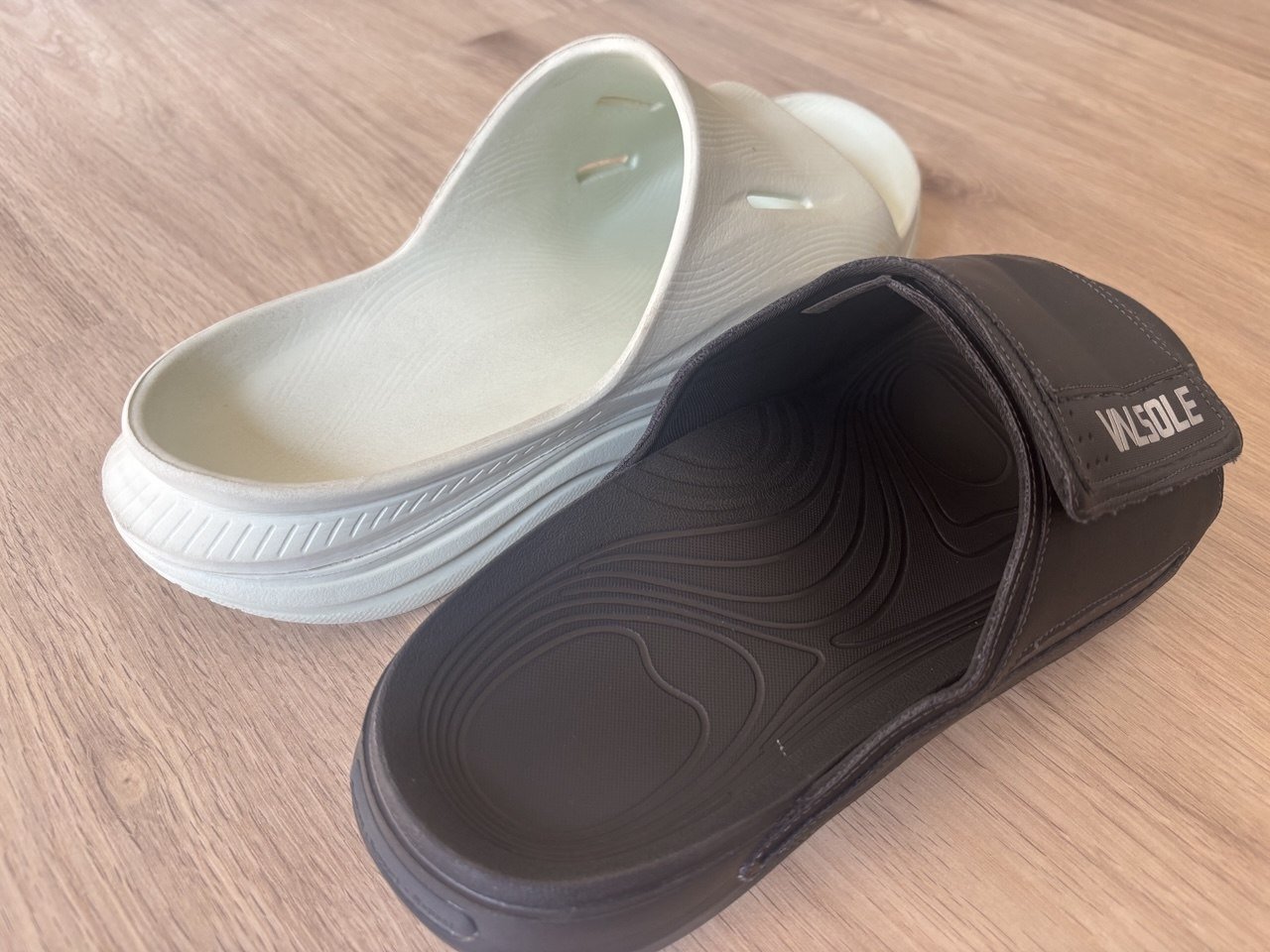Black VALSOLE Recovery Slide beside a white HOKA ORA Recovery Slide 3.